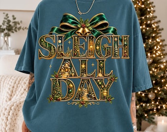 May include: A blue short-sleeve t-shirt featuring a large graphic print with the text "SLEIGH ALL DAY" in gold lettering. The design includes a green ribbon bow, golden bells, and detailed winter scenes with Christmas trees and lights.