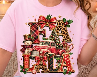 May include: A light pink t-shirt featuring a festive graphic with the text FA LA LA. The design includes a red vintage truck, a snowman in a top hat, a Christmas tree, red bows, and holly, with letters patterned in red plaid and leopard print.