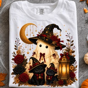 May include: A white t-shirt featuring a graphic of a ghost wearing a large black witch hat adorned with red roses and gold stars. Two black kittens wearing matching hats sit below the ghost next to a glowing lantern, a crescent moon, and roses.