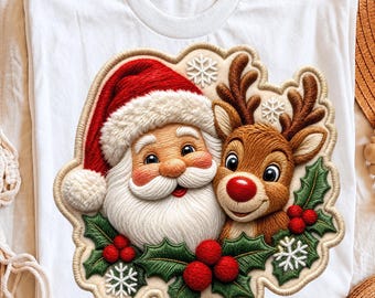 May include: A white t-shirt featuring a large, embroidered-style graphic of Santa Claus and a reindeer. The design includes red holly berries, green leaves, and white snowflakes, all framed by a scalloped, light-colored border.