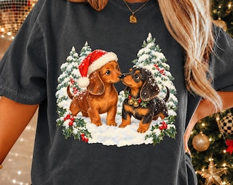 May include: A charcoal gray t-shirt featuring a graphic of two dachshunds in the snow. One dog wears a red Santa hat, and the other wears a green wreath collar with gold bells. They are surrounded by snow-covered pine trees and red holly berries.