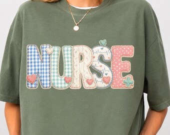 May include: An olive green short-sleeve t-shirt featuring the word NURSE in large, multi-patterned letters. Each letter has a unique design including blue gingham, green plaid, cream cable knit, medical EKG lines, and pink polka dots.