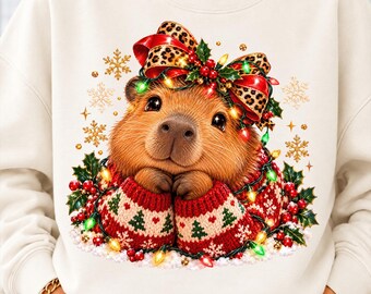 May include: A cream-colored sweatshirt featuring a graphic of a brown capybara wearing a red and white knit sweater with a Christmas tree pattern. The capybara has a leopard print bow with holly berries and glowing multicolored string lights.