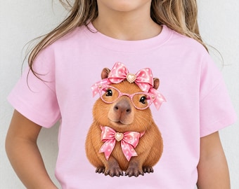 May include: A light pink short-sleeved t-shirt featuring a graphic of a brown capybara wearing pink polka-dot bows on its head and neck, along with pink cat-eye glasses. The capybara has a small heart-shaped embellishment on each bow.