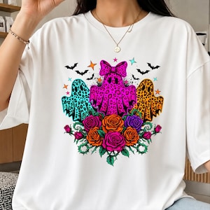 May include: A white short-sleeved t-shirt featuring a graphic print of three ghosts with leopard print patterns in teal, hot pink, and orange. The central pink ghost wears a bow. Below are colorful roses in pink and orange with green leaves.