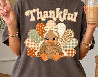 May include: A charcoal gray t-shirt featuring the word Thankful in a light-colored font above a graphic of a cartoon turkey. The turkey has a bow on its head and a tail made of various patterned feathers including polka dots, plaid, and leopard print.