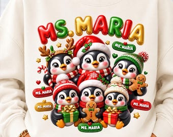 May include: A cream-colored sweatshirt featuring a graphic of six penguins wearing winter hats and scarves. The penguins hold gifts and gingerbread cookies. The text "MS. MARIA" appears in large, colorful, 3D-style letters and on small red and gold labels.