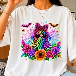 May include: A white t-shirt featuring a colorful graphic of a ghost with a leopard print pattern, wearing a matching leopard print bow and heart-shaped sunglasses. The design includes spider webs, bats, roses, and a sunflower in vibrant colors.