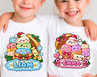 May include: Two white t-shirts featuring colorful cartoon illustrations of stacked dumplings in bamboo steamers. One shirt displays the name LIAM in blue, and the other displays the name EMMA in pink, surrounded by holiday motifs like snowflakes and bows.