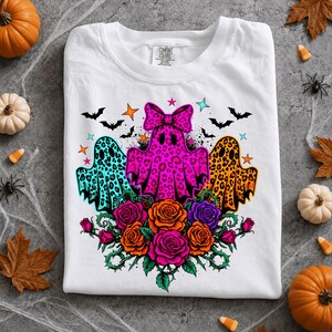 May include: A white t-shirt featuring a graphic of three ghosts with leopard print patterns in teal, hot pink, and orange. The center ghost wears a large pink bow. Below the ghosts are orange and purple roses with green vines and small bats.