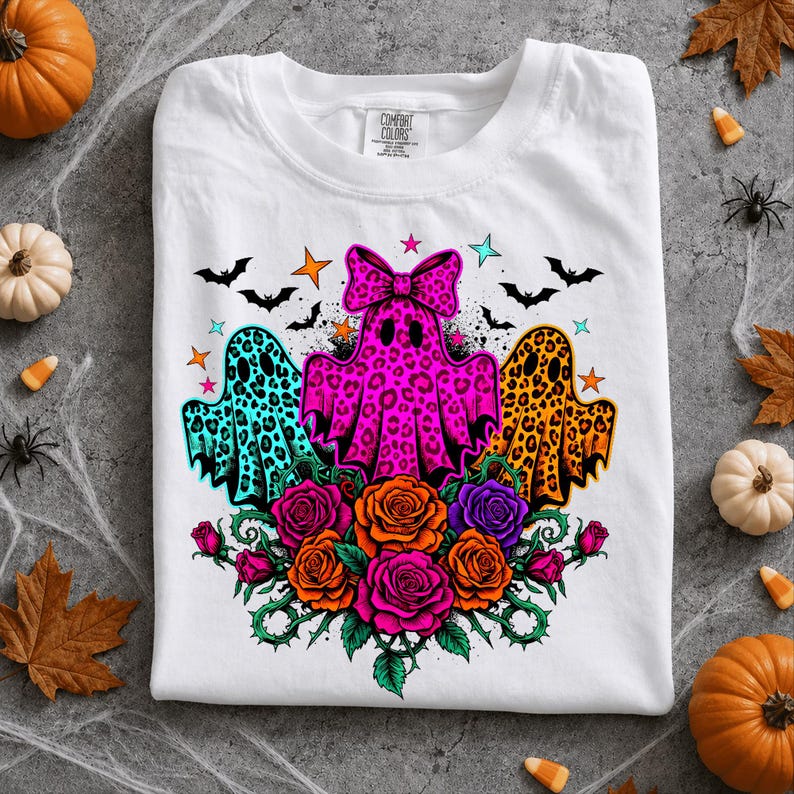 May include: A white t-shirt featuring a graphic of three ghosts with leopard print patterns in teal, hot pink, and orange. The center ghost wears a large pink bow. Below the ghosts are orange and purple roses with green vines and small bats.