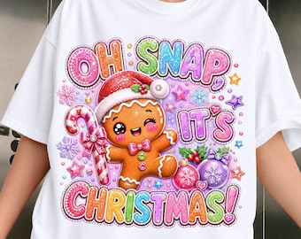 May include: A white short-sleeved t-shirt featuring a colorful graphic of a gingerbread person wearing a Santa hat, holding a candy cane. The text reads "OH SNAP, IT'S CHRISTMAS!" surrounded by glittery stars, snowflakes, and holiday ornaments.