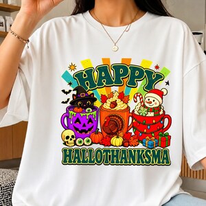 May include: A white t-shirt featuring a colorful graphic with the text HAPPY HALLOTHANKSMA. The design includes three mugs: a purple jack-o'-lantern mug with a black cat, an orange mug with a turkey, and a red mug with a snowman and gifts.