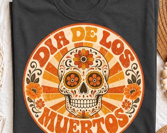 May include: A charcoal gray t-shirt featuring a circular graphic with a central sugar skull surrounded by orange and yellow flowers. The text "DIA DE LOS MUERTOS" is printed in a curved, retro-style font around the skull, set against a sunburst background.