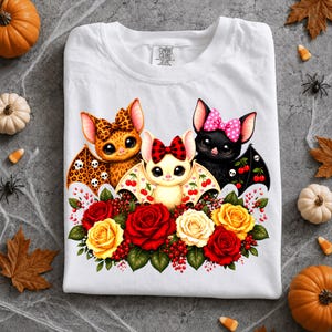 May include: A white t-shirt featuring a graphic of three cartoon bats with bows, surrounded by red and yellow roses. The left bat has a leopard print, the center bat is cream-colored with a plaid bow, and the right bat is black with a pink polka dot bow.
