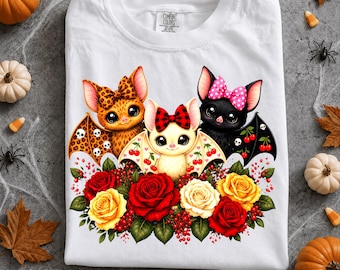 Puede incluir: Una camiseta blanca con un gráfico de tres murciélagos de dibujos animados con lazos, rodeados de rosas rojas y amarillas. El murciélago de la izquierda tiene estampado de leopardo, el central es color crema con un lazo de cuadros y el de la derecha es negro con un lazo rosa de lunares.