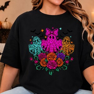 May include: A black t-shirt featuring a graphic print of three ghosts with leopard print patterns in turquoise, hot pink, and orange. The center ghost wears a bow. Below the ghosts are colorful roses in pink, orange, and purple with green stems.