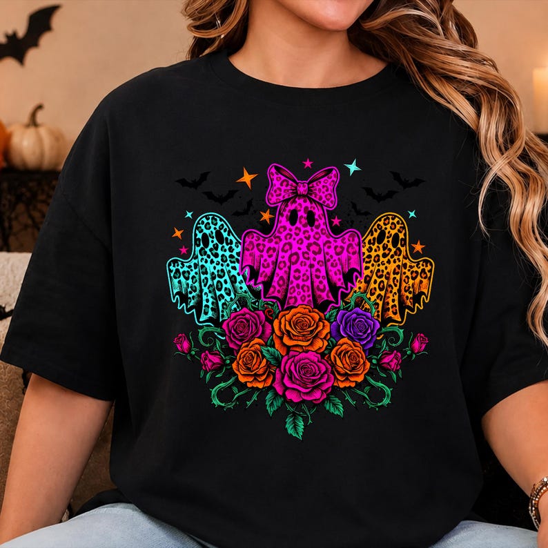 May include: A black t-shirt featuring a graphic print of three ghosts with leopard print patterns in turquoise, hot pink, and orange. The center ghost wears a bow. Below the ghosts are colorful roses in pink, orange, and purple with green stems.