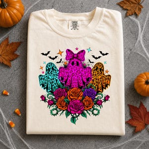 May include: A cream-colored t-shirt featuring a graphic of three ghosts with leopard print patterns in turquoise, hot pink, and orange. The center ghost wears a bow. Below the ghosts are colorful roses, and above are small bats and stars.