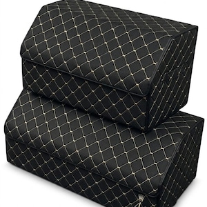 Puede incluir: Dos cajas de almacenamiento rectangulares negras con un patrón de diamantes acolchado y costuras doradas. Las cajas tienen tapas estructuradas y un logotipo de corona dorada con la letra N bordada en la esquina inferior de la caja.