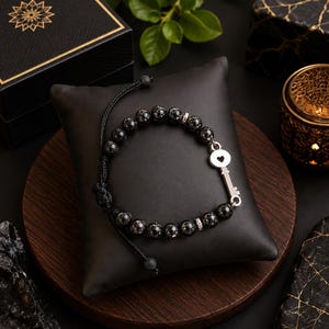 Peut inclure: Un bracelet de perles composé de pierres sombres et mouchetées, agrémenté d'une breloque en métal argenté en forme de clé avec une découpe en cœur. Le bracelet est doté d'un cordon noir avec un nœud coulissant réglable et des intercalaires argentés.