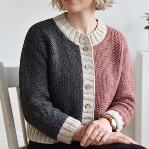 May include: A color-blocked knit cardigan featuring a dark charcoal gray right side and a dusty rose pink left side. The garment has a cream-colored ribbed crew neckline, a central button placket with four wooden buttons, and matching ribbed cuffs.