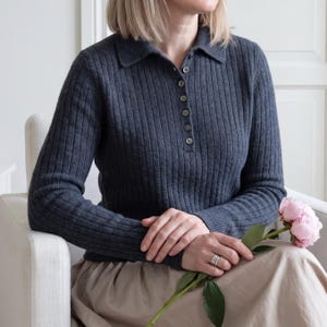May include: A dark charcoal grey long-sleeve polo shirt featuring a vertical ribbed knit texture, a classic collar, and a front placket with six small matching buttons. The garment is worn with a light beige skirt and a single pink peony flower.