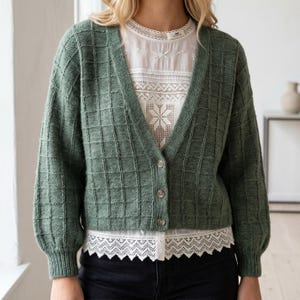 Peut inclure: Un gilet en tricot vert sauge avec un motif à carreaux texturé, une encolure en V et trois boutons sur le devant. Le gilet possède des manches longues avec des poignets côtelés et est porté sur un chemisier blanc avec des détails en dentelle.