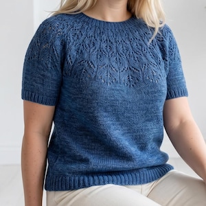 May include: A short-sleeved blue knit sweater featuring a round neckline and an intricate lace leaf pattern across the yoke and shoulders. The garment is constructed with a ribbed collar, ribbed sleeve cuffs, and a ribbed hem in a solid blue yarn.