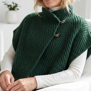 May include: A dark forest green knit poncho featuring a prominent chevron texture pattern, a high collar, and two brown wooden toggle buttons fastened on the front. The garment is worn over a plain white long-sleeved shirt with a high neckline.
