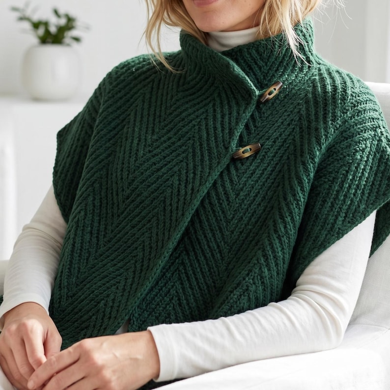 May include: A dark forest green knit poncho featuring a prominent chevron texture pattern, a high collar, and two brown wooden toggle buttons fastened on the front. The garment is worn over a plain white long-sleeved shirt with a high neckline.