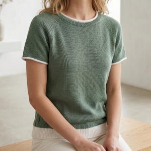 Puede incluir: Una prenda de punto de manga corta en color verde salvia apagado, con cuello redondo con borde interior blanco, puños blancos y dobladillo acanalado blanco. La prenda presenta mangas raglán y una textura de punto fina y uniforme.