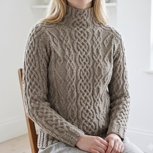 May include: A beige mock neck jumper featuring an intricate cable knit pattern with vertical braided designs across the front and sleeves. The garment is constructed with a ribbed collar, cuffs, and hem, showcasing a textured knit finish.