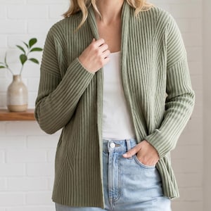 May include: A sage green open-front cardigan featuring a vertical ribbed knit texture, long sleeves, and a shawl collar. The garment is worn over a plain white top and light blue denim jeans, showcasing a consistent knit pattern throughout the piece.