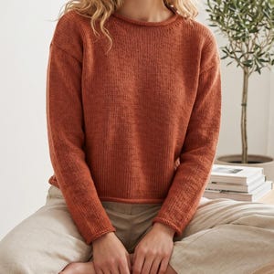 Op de afbeelding: Een trui met lange mouwen in een effen gebrande oranje kleur met een eenvoudige ronde halslijn met een opgerolde randafwerking. Het kledingstuk is geconstrueerd met een standaard breipatroon, lange mouwen en een rechte zoom op de taille.