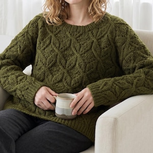 May include: An olive green long-sleeve jumper featuring a prominent cable knit pattern with a round neckline and ribbed cuffs. The jumper is paired with dark grey trousers and is held alongside a two-toned ceramic mug with a light grey base.