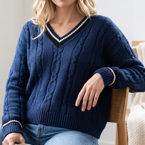 Peut inclure: Un pull en tricot torsadé bleu marine doté d'un col en V avec une bordure rayée noire et blanche. Ce modèle à manches longues présente un ourlet côtelé et des poignets rayés assortis, avec un motif de torsades verticales sur l'ensemble.