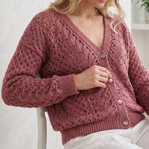 May include: A dusty rose-coloured knit cardigan featuring a V-neckline, long sleeves, and a delicate openwork lace pattern throughout. The garment includes a ribbed hem and cuffs, and is fastened down the front with four matching round buttons.