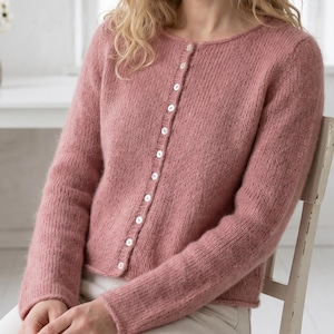 Könnte beinhalten: Eine altrosa Strickjacke mit Rundhalsausschnitt und einer vertikalen Reihe kleiner, weißer, runder Knöpfe an der Vorderseite. Das Kleidungsstück hat lange Ärmel und eine weiche, leicht flauschige Textur, die sich über das gesamte Strickgewebe erstreckt.
