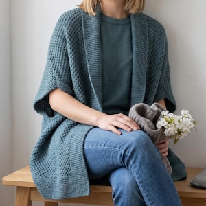 May include: A teal blue open-front cardigan featuring a textured waffle knit pattern, short sleeves, and a wide ribbed border. The garment is worn over a matching teal blue t-shirt and blue denim jeans, with a small bouquet of white flowers held.