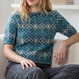 May include: A short-sleeved knit jumper featuring a teal and cream geometric Fair Isle pattern. The garment has a round neckline and ribbed trim at the collar, cuffs, and hem, showcasing a repeating intricate design across the entire front.