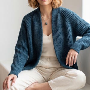 Könnte beinhalten: Eine tiefblaue, offene Strickjacke mit grober Rippstrickstruktur, langen Ärmeln mit gerippten Bündchen und einem Schalkragen. Das Kleidungsstück wird über einem cremefarbenen Unterhemd und einer hellen Hose getragen, ergänzt durch eine Silberkette.
