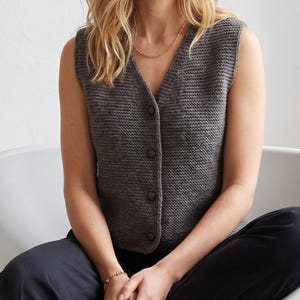 May include: A dark gray sleeveless knit vest featuring a V-neckline and a front button closure with four matching dark buttons. The garment is constructed with a textured, uniform stitch pattern and has a clean, sleeveless design.