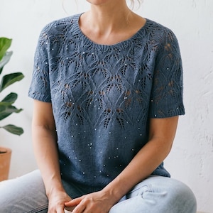 May include: A dusty blue short-sleeved knit top featuring an intricate lace leaf pattern across the upper chest and shoulders, accented with small white beads scattered throughout the design, paired with light blue denim jeans.