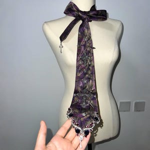 Pode incluir: Uma gravata com padrão roxo e preto com um laço no colarinho, pingentes de cruz prateados, espigões de metal e uma extremidade pontiaguda adornada com pedras escuras em forma de lágrima e cristais transparentes, num manequim.