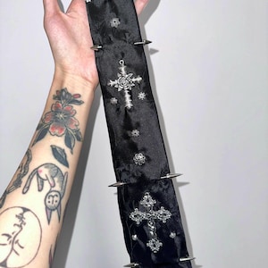 Peut inclure: Une cravate en satin noir avec un motif floral jacquard subtil, ornée de deux pendentifs en forme de croix argentée, de plusieurs petits clous floraux argentés et de quatre pointes coniques argentées sur les bords.