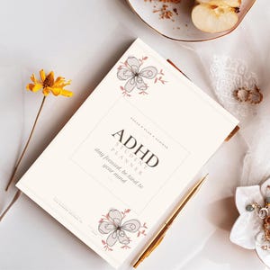 Peut inclure: Un agenda étudiant TDAH de couleur crème avec une bordure rectangulaire, des dessins floraux aux coins et un texte noir indiquant ADHD STUDENT PLANNER, FOCUS & PLAN & ACHIEVE, et stay focused, be kind to your mind, avec un stylo doré.