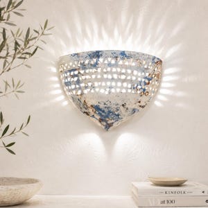 Può includere: Un'applique da parete semicircolare con finitura bianca e blu anticata e intagli triangolari. La lampada emette luce attraverso le aperture, proiettando un motivo a raggiera sulla parete, e illumina verso il basso dalla sua base.