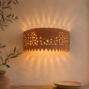 Può includere: Un'applique semicircolare in terracotta con intricati ritagli triangolari lungo il bordo superiore e un bordo inferiore seghettato. L'apparecchio emette una luce calda che proietta raggi distinti sulla superficie della parete.