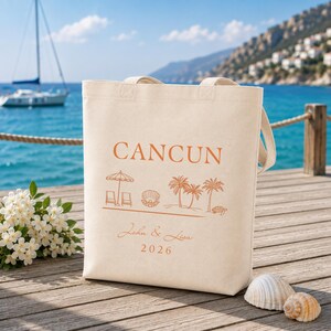 May include: A beige canvas tote bag featuring a printed design in orange ink. The text reads CANCUN, John & Lisa, and 2026. The graphic includes a beach umbrella, two chairs, a seashell, palm trees, and a sea turtle on a horizontal line.